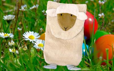 Die plüschige Wärmflasche Lilo Lamm sitzt in einer Wiese, man sieht bunte Ostereier