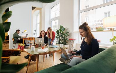 Eine Frau im Vordergrund sitzt auf dem Sofa und arbeitet am Laptop, der auf ihrem Schoß liegt. Im Hintergrund sieht man drei weitere Personen, die in der Arbeit vertieft sind. 