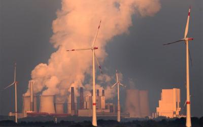 Windkraftwerke im Vordergrund, im Hintergrund befindet sich ein Kraftwerk, dass viel Rauch ausstoßt.
