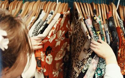 Angeschnitten ist ein Kopf von einer jungen Frau von Hinten. Sie zieht mit ihren Händen bunte Hemden auseinander, die auf einer Kleiderstange hängen.