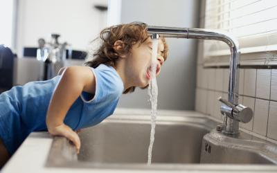 Kleines Kind mit braunen Haaren und blauem T-Shirt beugt sich über die Küchenzeile um aus dem Wasserhahn zu trinken.