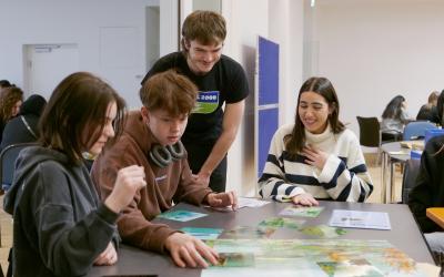 Ein junger Mann mit GLOBAL 2000 T-Shirt beugt sich über einen Tisch, an dem drei Schüler:innen sitzen und an einer Aufgabe arbeiten. Alle lachen.