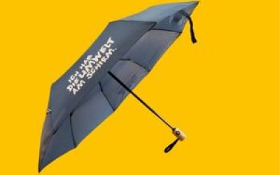 Ein dunkelblauer Klappschirm mit Holzgriff ist vor einem leuchtend gelben Hintergrund geöffnet. Der weiße Text auf dem Schirm lautet: „Ich habe die Umwelt am Schirm."