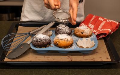 Eine Person streut Staubzucker auf Muffins, die sich noch in der Backform befinden.