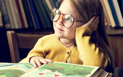 Ein Mädchen im gelben Pulli und mit großer, runder Brille sitzt vor einem Märchenbuch schaut verträumt zur Seite.