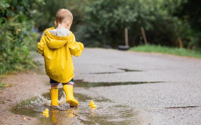 Kleinkind mit gelber Regenjacke und gelben Gummistiefel spielt mit Gummienten in einer Pfütze