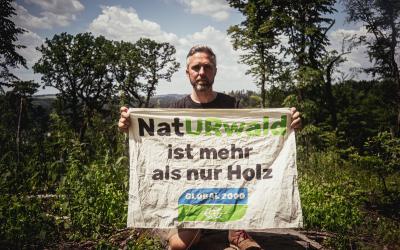 Dominik Linhard mit einem Banner im Wald: "NatURwald ist mehr als nur Holz"