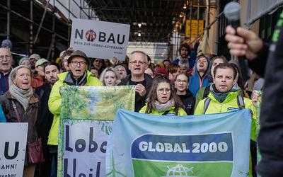 ine große Gruppe von Demonstrierenden steht dicht gedrängt unter einem Gerüst. Vorne halten mehrere Menschen Banner hoch, darunter eines mit der Aufschrift ‚GLOBAL 2000‘ und ein anderes mit ‚Lobau bleibt‘. Im Hintergrund sind weitere Protestschilder zu sehen, etwa mit ‚No Bau – Nein zur Lobau-Autobahn‘. Die Stimmung wirkt entschlossen und kämpferisch.