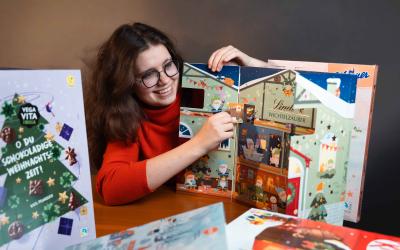 Mädchen in rotem Pulli öffnet Fenster von Schoko-Adventkalender, rundherum mehrere Adventkalender