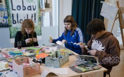 Drei Personen sitzen an einem Tisch, der mit Bastelmaterialien bedeckt ist, und fertigen Kunstwerke oder Plakate an. Bunte Papiere, Marker und Körbe liegen auf dem Tisch verstreut. Im Hintergrund ist ein Schild zu sehen, auf dem „Lobau bleibt!“ steht.