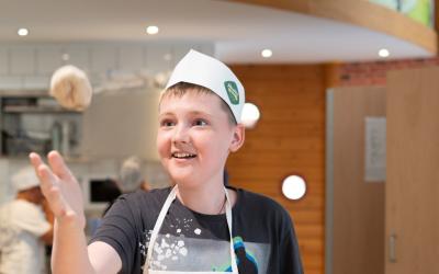 Junge mit Schürze und Kopfbedeckung schupft lachend Teig beim backen.