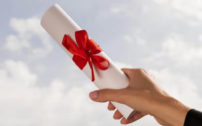 Hand hält eine zusammengerollte weiße Urkunde mit roter Schleife vor bewölktem Himmel