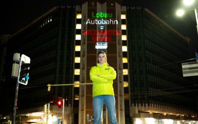 In der Mitte des Bildes steht eine junge Frau mit entschlossenem Blick und verschränkten Armen vor einem Gebäude, dem österreichischen Verkehrsministerium. Die Frau trägt eine Jacke mit einem Global 2000 Logo. Auf das Gebäude hinter ihr ist ein Schriftzug gebeamt "Lobau Autobahn = Milliardengrab!"