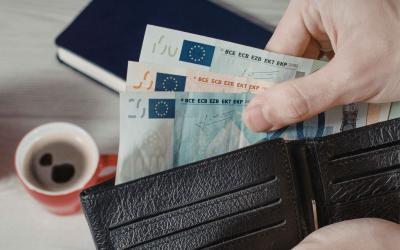 Eine Hand zieht mehrere Euro Geldscheine aus einer Geldbörse, auf dem Tisch dahinter steht ein Kaffee und es liegt ein Notizbuch daneben