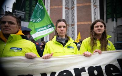 Mehrere Personen in knallgelben Jacken und GLOBAL 2000-Logo mit Banner in den Händen, dahinter Fahnen, auf einer Demo
