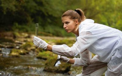 Wissenschaftlerin in weißem Schutzanzug und Handschuhen entnimmt Wasserprobe aus einem Fluss