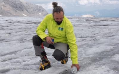 Person in gelber Jacke kniet auf einem Gletscher und hält eine Wasserflasche auf dem Eis