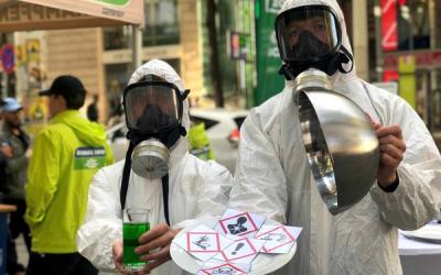 Zwei Personen in weißen Schutzanzügen und Atemschutzmasken halten ein Glas mit grünem Flüssigkeit und ein rundes Metallobjekt, davor ein Papier mit Gefahrensymbolen.