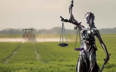 Statue der Justitia mit Waage im Vordergrund vor einem Traktor, der auf einem Feld Pestizide versprüht