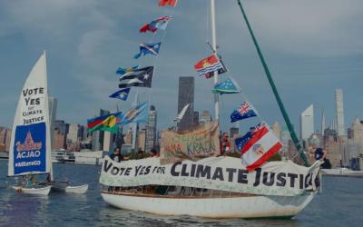 Segelboot mit mehreren Flaggen und Transparenten mit der Aufschrift 'Vote Yes for Climate Justice' vor einer Stadtsilhouette