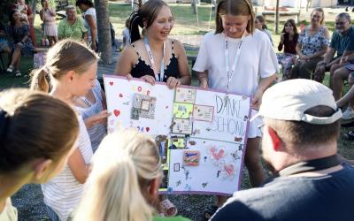 Drei junge Frauen zeigen im Freien ein großes, bunt gestaltetes Plakat mit der Aufschrift 'DANKE SCHÖN' vor einer Gruppe sitzender und stehender Menschen.