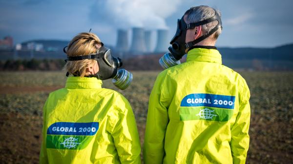 Zwei GLOBAL 2000 Aktivist:innen mit Gasmasken stehen vor einem Atomkraftwerk