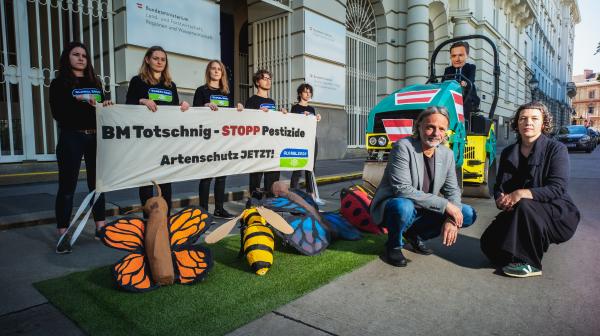 Totschnig fährt mit einer Walze über Bestäuber. Im Hintergrund stehen Aktivist:innen mit einem Plakat.