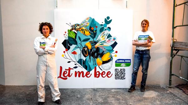 zwei Aktivistinnen mit Pinsel in den Händen stehen vor dem Bild, dass sie gerade gemalt haben. Darauf zu sehen sind zwei Bienen und der Schriftzug "Let me bee"
