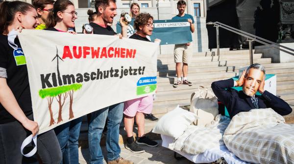 Aktivist:innen halten Banner in der Hand, worauf steht: "Aufwachen: Klimaschutz jetzt!". Ein Aktivist mit einer Nehammer-Maske liegt daneben im Bett und hält sich die Ohren zu.