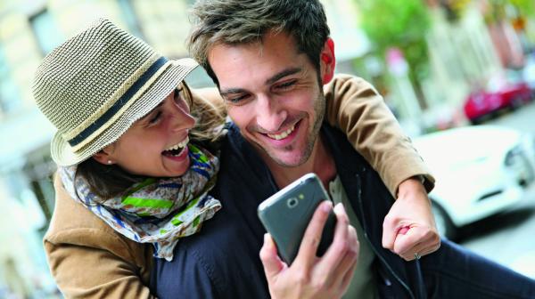 Eine Frau mit Hut umarmt einen Mann und beide schauen freudig in ein Smartphone.