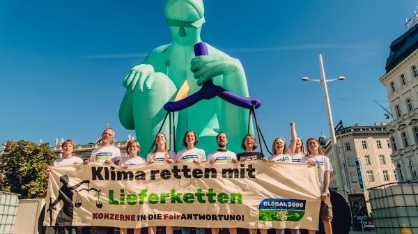 Aktivist:innen halten einen Banner in der Hand mit der Aufschrift "Klimaretten mit Lieferketten". Sie stehen vor einer 10 Meter großen Statue, die Justitia darstellt.