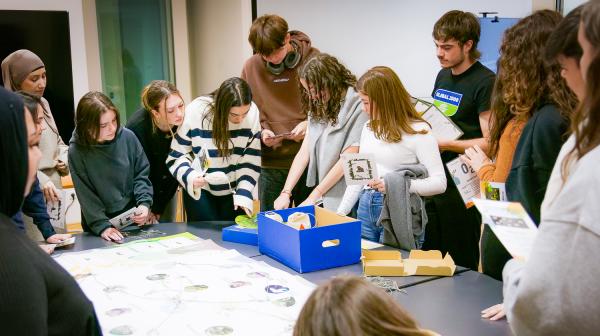 Schülergruppe steht um einen Tisch und sie schauen interessiert in eine blaue Box. Einige von ihnen halten eine Karte in den Händen. 