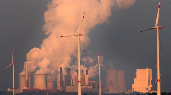 Windkraftwerke im Vordergrund, im Hintergrund befindet sich ein Kraftwerk, dass viel Rauch ausstoßt.