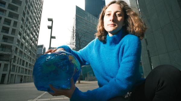 Anna Leitner trägt einen blauen Pullover, sie kniet am Boden vor hohen Bürogebäude und hält eine Plastik-Weltkugel in der Hand
