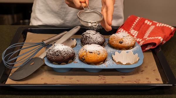Eine Person streut Staubzucker auf Muffins, die sich noch in der Backform befinden.
