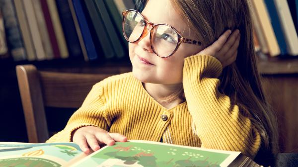 Ein Mädchen im gelben Pulli und mit großer, runder Brille sitzt vor einem Märchenbuch schaut verträumt zur Seite.