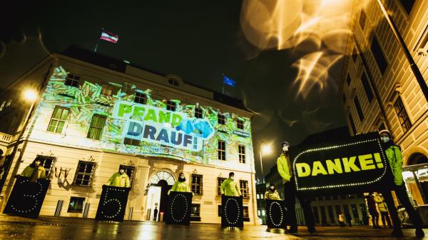 Beamer-Aktion mit dem Pfand-drauf-Logo. Davor stehen Aktivist*innen mit einem Banner, auf dem "Danke" steht.