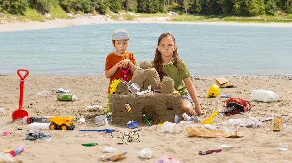 2 Kinder bauen Sandburg mit Müll drin, schauen traurig