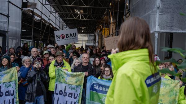 Protestierende vor dem Verkehrsministerium in Wien. Einige tragen Jacken mit dem Logo von GLOBAL 2000, viele halten Banner mit der Aufschrift "Lobau bleibt!" oder "NOBAU". Eine Sprecherin von GLOBAL 2000 steht im Vordergrund und spricht zur Menge.