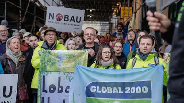 ine große Gruppe von Demonstrierenden steht dicht gedrängt unter einem Gerüst. Vorne halten mehrere Menschen Banner hoch, darunter eines mit der Aufschrift ‚GLOBAL 2000‘ und ein anderes mit ‚Lobau bleibt‘. Im Hintergrund sind weitere Protestschilder zu sehen, etwa mit ‚No Bau – Nein zur Lobau-Autobahn‘. Die Stimmung wirkt entschlossen und kämpferisch.