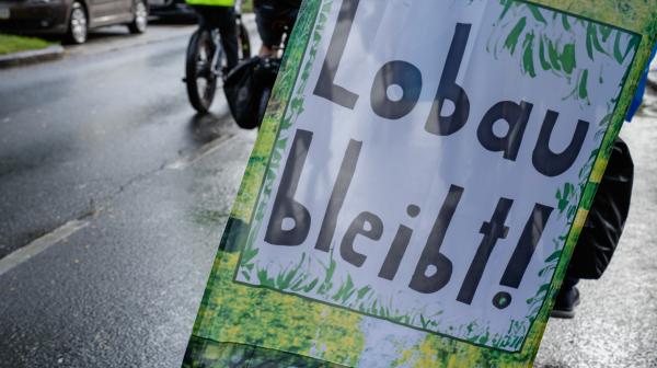Banner mit der Aufschrift "Lobaut bleibt!" hängt an einem Fahrrad, das auf einer nassen Straße fährt.