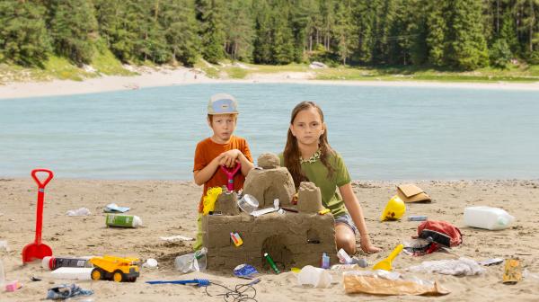2 Kinder bauen eine Sandburg mit Müll vor österreichischer Landschaft