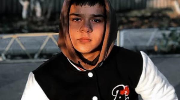 Junge mit schwarzen Haaren und Schwartz weißer Jacke sitzt auf einer Bank 