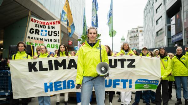 Viele Menschen auf einer Demonstration. Im Vordergrund steht eine junge Frau in GLOBAL 2000 Jacke mit einem Megafon. Im Hintergrund sind Banner mit der Aufschrift "Klimaschutz statt heißer Luft" und "Autobahnen sind ein Graus bauen wir die Öffis aus". Viele Menschen im Hintergrund tragen Fahnen und Jacken von GLOBAL 2000.