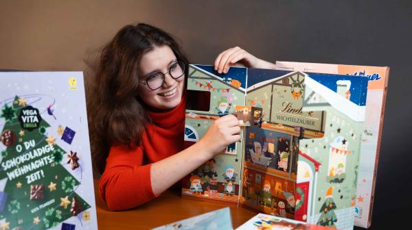 Mädchen in rotem Pulli öffnet Fenster von Schoko-Adventkalender, rundherum mehrere Adventkalender