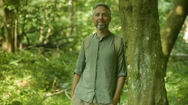 Ein Mann steht in einem grünen Wald an einen Baum gelehnt. Er ist zentral im Bild zu sehen, mit den Händen in den Hosentaschen und Blick in die Kamera.