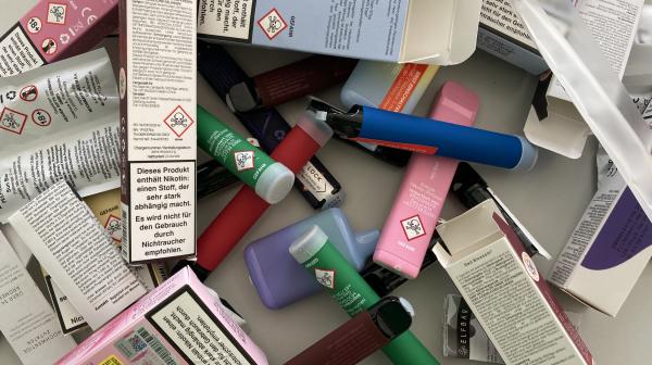Einweg Vapes mit und ohne Verpackung auf einem Haufen. Überall sind Warnhinweise und Totenköpfe auf den Produkten und den Verpackungen zu sehen.