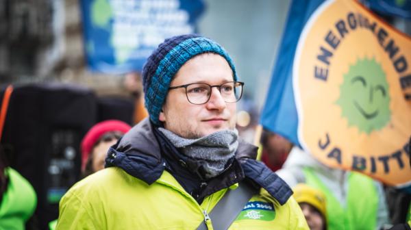 Ein Mann mit Brille und Haube in einer GLOBAL 2000 Jacke steht vor einer Menschengruppe. Im Hintergrund wird eine Fahne hochgehalten mit der Aufschrift "Energiewende Ja bitte"