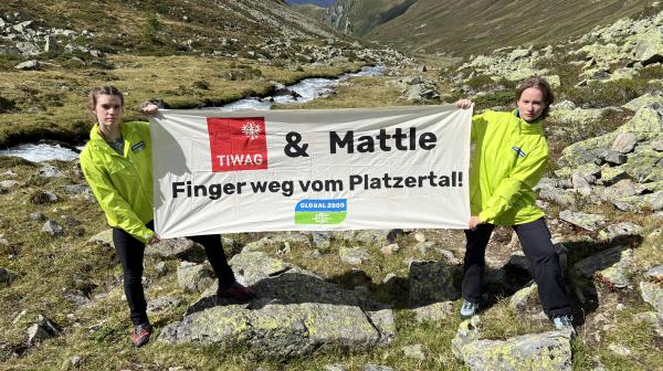 Zwei Personen mit GLOBAL 2000 Jacken stehen in einer Berlandschaft, im Hintergrund ist ein Bach zu sehen. Sie halten ein Banner mit der Aufschrift "TIWAG & Mattle - Finger weg vom Platzertal!"