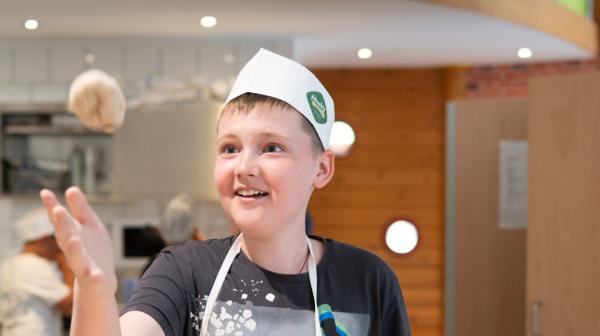 Junge mit Schürze und Kopfbedeckung schupft lachend Teig beim backen.